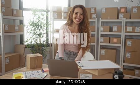 Frau, die auf einem Laptop schreibt, während sie Paketetiketten an einem Schreibtisch im Gebäude überprüft; Produktivität kleiner Unternehmen. Stockfoto