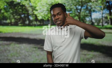 Mann mit weißem Polohemd und Brille von Hand zu Ohr, der im Wald unter sonnendurchflutetem Baldachin Telefongeste macht; Kommunikationsfreude. Stockfoto