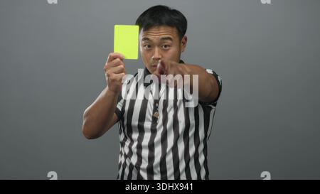 Junger chinesischer Schiedsrichter hält gelbe Karte und zeigt mit dem Finger, während er ein gestreiftes Hemd im grauen Studio trägt; Autorität. Stockfoto