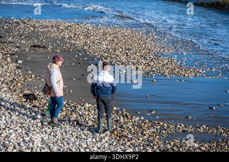 Junge Paare stehen zusammen an einem Kieselstrand oder Steinstrand bei Ebbe und beobachten das Wasser und die Wellen. Stockfoto