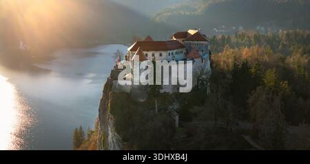 Panoramablick auf Schloss Bled auf einer steilen Klippe bei Sonnenuntergang. Sanftes goldenes Stundenlicht, das die mittelalterliche Festung mit warmem Glanz und schöner Schönheit beleuchtet Stockfoto
