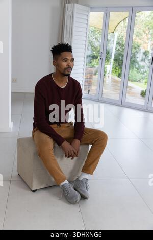 Mann mit kastanienbraunem Pullover, der auf Ottomane in einem hellen Wohnzimmer in der Nähe von Schiebetüren und Rollläden sitzt Stockfoto