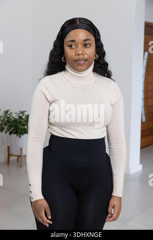Erwachsene afroamerikanische Frau posiert im Foyer in beigem Pullover, schwarzer Hose, Topfpflanze, Holztür Stockfoto
