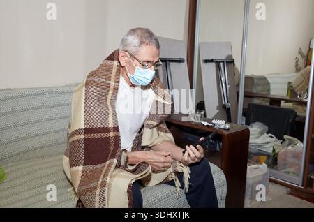 Ein älterer Mann ist zu Hause krank. Ein alter Mann sitzt in einem Karo, trägt eine Hygienemaske und hält ein Smartphone in der Hand. Patient isoliert auf Stockfoto