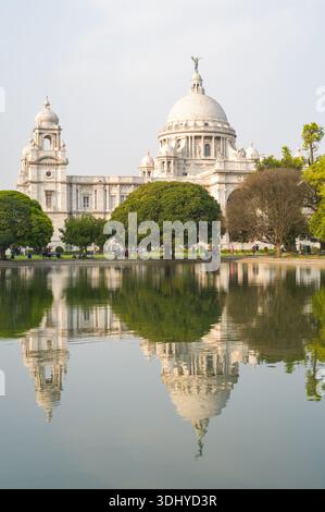 Calcutta, Indien - 23. März 2024: Das Bild zeigt das Victoria Memorial mit seiner Marmorkuppel und Türmen, die sich in einem großen Wasserbecken spiegeln, umgeben von grünen Bäumen und Menschen im Garten unter klarem Himmel. Stockfoto