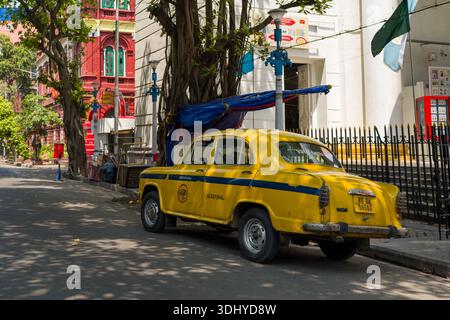 Calcutta, Indien - 24. März 2024: Ein gelbes Ambassador-Taxi parkt in einer ruhigen Straße neben einem weißen Gebäude mit Säulen, schwarzem Eisenzaun und Beschilderung, mit einem roten Gebäude aus der Kolonialzeit und grünen Bäumen, die in Kalkutta zu sehen sind. Stockfoto