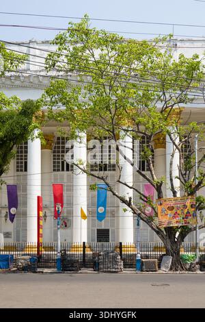 Calcutta, Indien - 24. März 2024: Hinter einem schwarzen Eisenzaun und Straßenlaternen in Kalkutta ist die Vorderseite des General Post Office Gebäudes mit weißen Säulen, bunten Bannern und einem großen Baum mit grünen Blättern zu sehen. Stockfoto