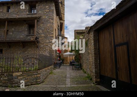 Unbekannte Lage - 6. Mai 2024: Blick auf eine enge kopfsteingepflasterte Gasse mit Cafétischen im Freien, einem großen weißen Sonnenschirm, Restaurantschildern und historischen Steinhäusern mit Balkonen und Holztüren. Stockfoto