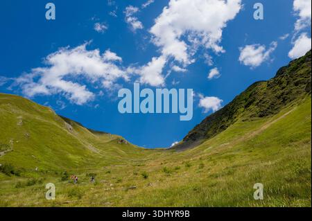 Col du tricot, Frankreich - 24. August 2024: Mehrere Wanderer sind zu sehen, die sich auf dem Weg durch ein weites grasbewachsenes Tal zwischen steilen grünen Hängen unter blauem Himmel mit verstreuten Wolken aufmachen. Stockfoto