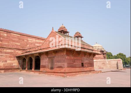 Fatehpur Sikri, Indien - 12. März 2024: Das Bild zeigt das Nebengebäude des Jodha Bai Palace mit roten Sandsteinwänden, einem schrägen Dach, einer dekorativen Attika und zwei kleinen Kuppelpavillons mit einem gepflasterten offenen Innenhof und Bäumen im Hintergrund unter klarem Himmel. Stockfoto