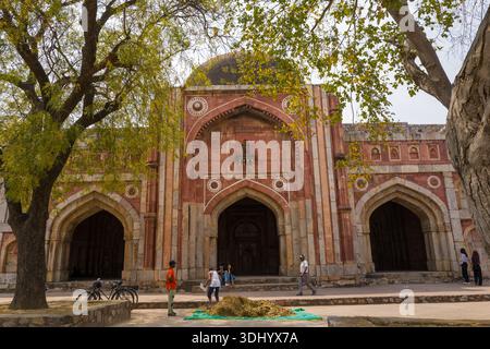 Delhi, Indien - 30. März 2024: Das Bild zeigt mehrere Menschen, die in der Nähe des bogenförmigen Haupteingangs der Jamali Kamali Moschee im Archäologischen Komplex Mehrauli laufen und stehen, mit großen Bäumen, die den Vorderboden umrahmen und Fahrrädern, die an der Seite geparkt sind. Stockfoto