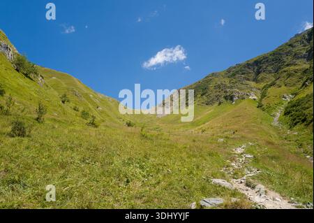 Col du tricot, Frankreich - 24. August 2024: Ein felsiger Wanderweg führt sich durch ein weites grasbewachsenes Bergtal zwischen steilen grünen Hängen unter klarem blauem Himmel mit einigen verstreuten Wolken. Stockfoto