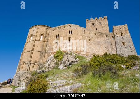 Loarre, Spanien - 8. Mai 2024: Die Apsis, die Steinmauern und die Türme der Burg Loarre sind auf einem felsigen Hügel mit grünem Gras, Sträuchern und einer Gruppe von Menschen am Fuß unter einem klaren blauen Himmel zu sehen. Stockfoto