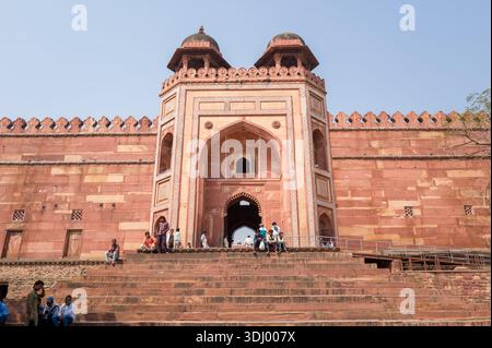 Fatehpur Sikri, Indien - 12. März 2024: Das Bild zeigt das sekundäre Eingangstor der Jama-Masjid-Moschee mit seiner roten Sandsteinfassade, dem bogenförmigen Portal und zwei Kuppelpavillons, in denen Menschen auf den Stufen vor dem Gebäude sitzen und stehen. Stockfoto