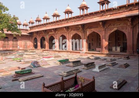 Fatehpur Sikri, Indien - 12. März 2024: Das Bild zeigt den Friedhofshof der Jama Masjid Moschee mit mehreren Gräbern, Gebetsteppichen, die einige Gräber bedecken, einen großen Baum auf der linken Seite und die rote Sandsteinfassade mit gewölbten Chhatris im Hintergrund, da einige Menschen in der Nähe der Bögen zu sehen sind. Stockfoto