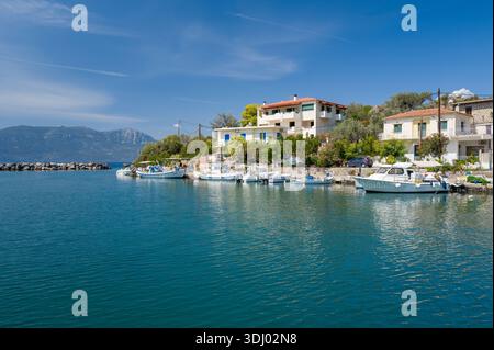 Port de Vathi, Griechenland - 3. September 2024: Reihe von kleinen Fischerbooten, die entlang der Uferpromenade vor Wohnhäusern vertäut sind, mit Bergen im Hintergrund unter einem klaren blauen Himmel. Stockfoto