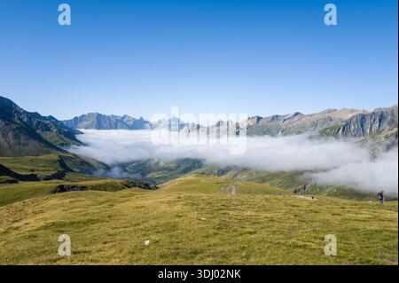 Frankreich - 26. August 2024: Eine Schicht niedriger Wolken füllt ein weites Bergtal unter einem klaren blauen Himmel, mit grasbewachsenen Hängen im Vordergrund und mehreren Wanderern am Hang. Stockfoto