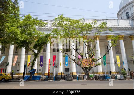 Calcutta, Indien - 24. März 2024: Hinter einem schwarzen Eisenzaun und Straßendrähten befindet sich die Vorderseite des General Post Office Gebäudes mit hohen weißen Säulen, goldenen Kapitellen, bunten Bannern und Laubbäumen. Stockfoto