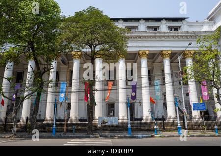 Calcutta, Indien - 24. März 2024: Die Fassade des General Post Office Gebäudes mit hohen weißen Säulen, goldenen Kapitellen, bunten Bannern, Laubbäumen und einem schwarzen Eisenzaun ist entlang einer Straße in Calcutta zu sehen. Stockfoto