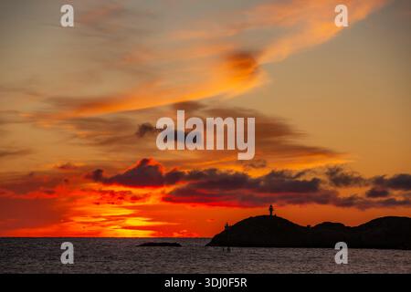 Svolvear, Lofoten Island, Norwegen wunderschöner Sonnenuntergang über dem Meer mit einem Leuchtturm in der Ferne. Der Himmel ist voller Wolken und die Sonne ist setti Stockfoto