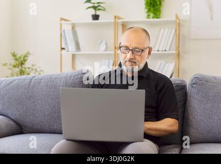 Seriöser älterer Mann, der eine Brille trägt, mit dem Laptop auf einem bequemen Sofa zu Hause Stockfoto