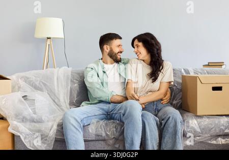 Ein lächelndes Paar umarmt sich auf dem Sofa zwischen Kisten, nachdem er in sein neues Zuhause gezogen ist Stockfoto