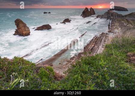 Weitwinkelblick auf eine zerklüftete Küste bei Sonnenuntergang mit dramatischen Meeresstapeln, Flysch-Felsschichten und stürzenden Meereswellen unter einem bunten Himmel. Stockfoto
