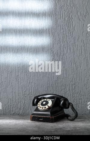 Vintage-schwarzes Telefon mit Drehknopf auf grauem Steintisch. Ein Lichtstrahl fällt durch ein Fenster mit Gitter auf die graue Wand. Das Konzept von HO Stockfoto