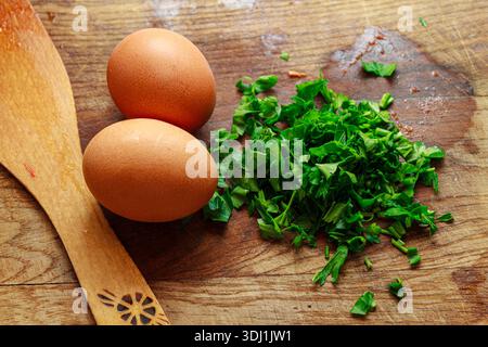 Zwei rohe braune Hühnereier und gehackte grüne Petersilie auf einem rustikalen Holzbrett. Zutaten zum Frühstück. Stockfoto