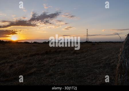 Ein dramatischer Sonnenuntergang über einem geernteten Feld mit runden Heuballen und einem Stromleitungsturm am Horizont unter einem goldenen Himmel. Stockfoto