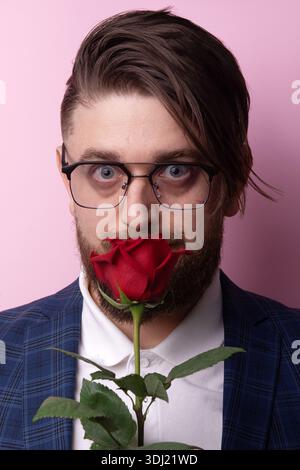 Bärtiger Mann in Brille mit roter Rose vor dem Gesicht, mit blauer Anzugsjacke und weißem Hemd, vor pastellrosa Hintergrund. Stockfoto