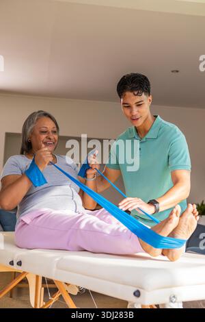 Leitende Afroamerikanerin und asiatische Therapeutin, die in der Klinik Blue-Band-Übungen auf dem Tisch durchführen. Rehabilitation, Physiotherapie, Wellness, Seniorengesundheit, Krafttraining, therapeutisch, Gesundheitswesen || Modell veröffentlicht Stockfoto