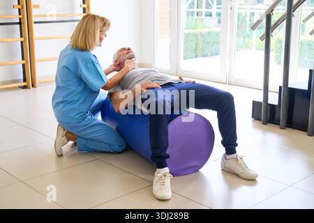 Ein älterer Mann liegt auf einem Trainingsball, während ein Physiotherapeut in einem Rehabilitationszentrum Nackentherapie anbietet. Diese Indoor-Sitzung konzentriert sich auf Rehabilitation und manuelle Therapie zur Erholung und Verbesserung des Wohlbefindens. || Modell freigegeben Stockfoto