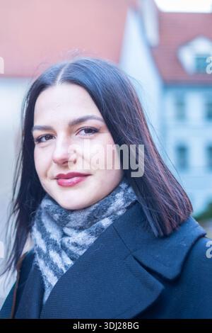 Eine junge Frau mit dunklen Haaren und einem Schal steht draußen in einer Stadt, mit verschwommenen Gebäuden, die einen weichen Hintergrundeffekt erzeugen Stockfoto