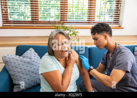 Ältere Frau zuzwinkernd, während asiatischer Gesundheitsdienstleister Spritze auf blauem Sofa im Wohnzimmer benutzt. Alten, Pflegepersonal, Medizin, Komfort, Wellness, Home Interior, professionell || Modell veröffentlicht Stockfoto