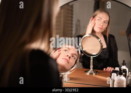 Eine junge Frau mit langen Haaren wendet Hautpflege auf ihr Gesicht an, während sie in einen runden Spiegel auf einem hölzernen Waschtisch blickt Stockfoto