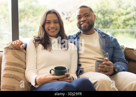 Verschiedene Paare lächeln, während sie Keramikbecher auf dem gepolsterten Sofa im Wohnzimmer am Fenster halten. Gemütlichkeit, Verbindung, Entspannung, Haushalt, Komfort, Harmony, modern || Modell veröffentlicht Stockfoto