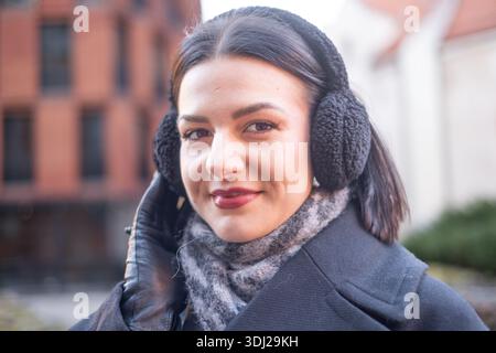 Eine junge Frau mit dunklen Haaren und Ohrenschützern lächelt im Freien in einer städtischen Umgebung, mit Ziegelhäusern und Grünanlagen im Stockfoto