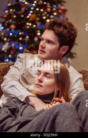 Das junge Paar entspannt sich auf einer Couch, die Frau lehnt sich an den Mann, während ein wunderschön geschmückter Weihnachtsbaum mit Lichtern steht Stockfoto