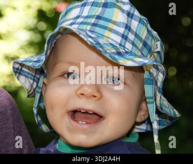 Lächelnder Junge im Freien mit kariertem Sonnenhut, Nahaufnahme-Porträt mit grünem Bokeh-Hintergrund Stockfoto
