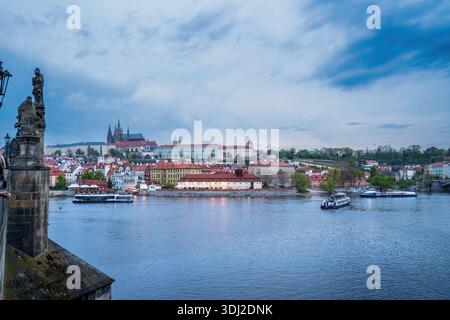 Eine ruhige Abendszene von Prag über die Moldau, mit farbenfrohen Gebäuden am Fluss, einer von Statuen gesäumten Brücke und Booten, die als C vorbeigleiten Stockfoto