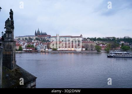 Ein Panoramablick auf Prag über die Moldau mit historischen Gebäuden, einer Burg auf einem Hügel, Statuen auf der Brücke und einem Boot, das entlang der Moldau fährt Stockfoto