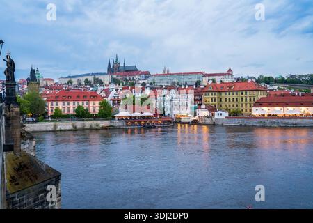 Ein malerischer Abendblick auf Prag über die Moldau mit farbenfrohen historischen Häusern, roten Dächern und einer fernen Burg. Boote leuchten mit Lichtern r Stockfoto