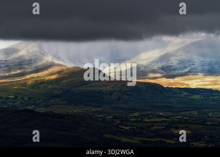 Die Sonne scheint durch die Wolken auf die grünen und braunen Hügel des Killarney National Park in Irland. Die fernen Berge sind bedeckt Stockfoto