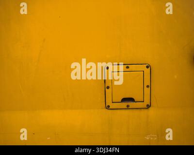 Minimalistische Nahaufnahme einer verwitterten gelben Industrietafel mit einer kleinen quadratischen Zugangsklappe und Schrauben, die authentische mechanische Textur zeigen Stockfoto