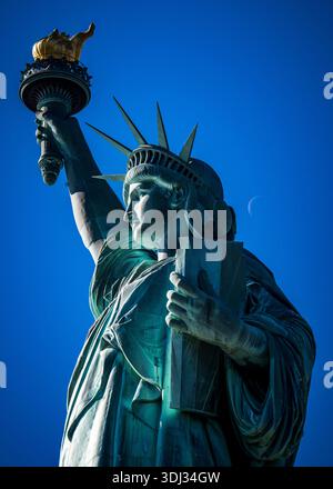 Freiheitsstatue aus nächster Nähe mit sichtbarem Mond hinter Fackel vor tiefblauem Himmel, Liberty Island, New York City. Stockfoto