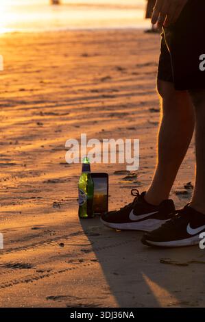 Eine Person steht am Sonnenuntergang in Tofino, Vancouver Island, mit schwarzen Sneakers und Shorts auf dem Sand. Eine grüne Peroni-Bierflasche und ein Smartphone sind es Stockfoto