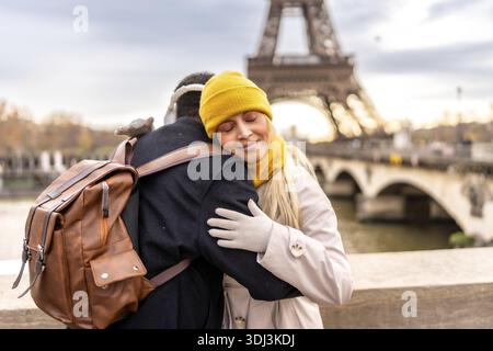 Junge, vielfältige Paare umarmt sich auf einer pariser Brücke und teilt einen zärtlichen Moment mit dem eiffelturm und seine Waden, die sanft im winterlichen Stadtbild verschwimmen Stockfoto