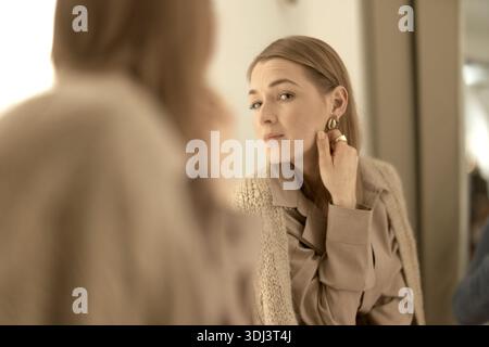 Eine elegante Frau, die in den Spiegel schaut und ihr Aussehen überprüft, ein beigefarbenes Outfit, ein sanftes Lächeln und elegante Ohrringe trägt, selbstbewusst, selbstverliebt Stockfoto