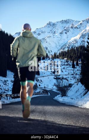 Mann Athlet, der auf der Straße im schneebedeckten Bergtal läuft, am sonnigen Wintertag, außer Fokus. Joggen in Sportbekleidung in der Natur. Stockfoto